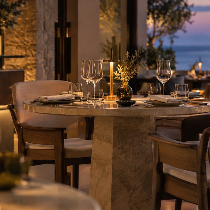 One laid setting at close range, glassware, a brass table lamp and an olive sprig on pale stone